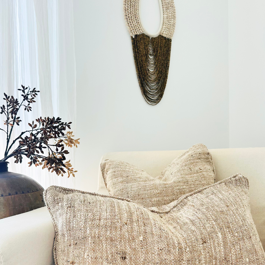 Two handwoven cushion covers in beige and brown tones on a sofa, with a large cowrie shell wall hanging above. A decorative plant and a framed artwork are also visible in the background.