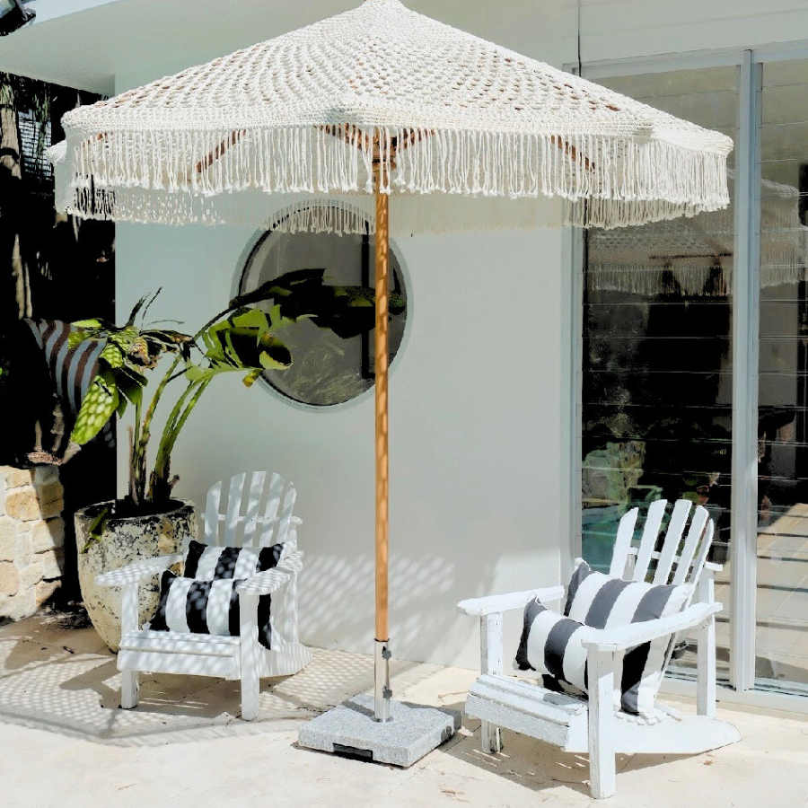 White patio umbrella with fringe and wooden pole over two white chairs with striped cushions by a poolside.