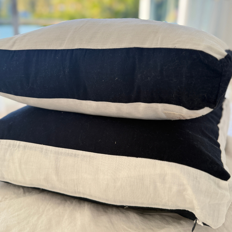 Two striped cushion, one navy and one beige, stacked on a surface with a blurred outdoor background.
