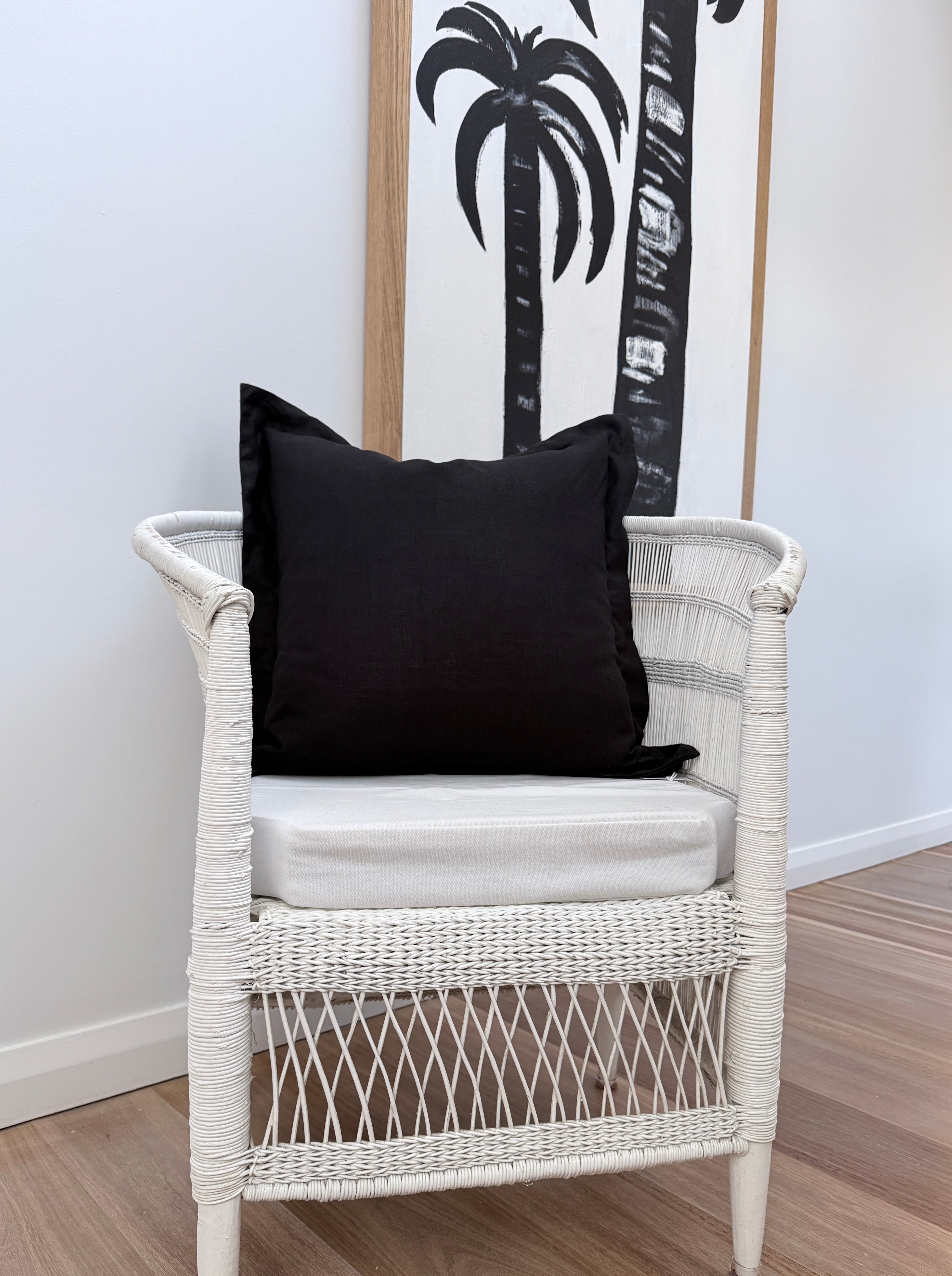 A white woven rattan armchair with a white cushion, styled with a black throw cushion, set against a white wall with a framed palm tree print.