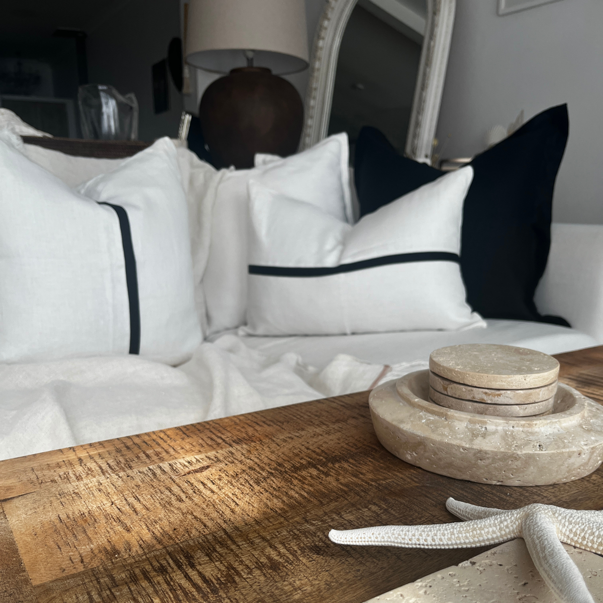 Ivory and black linen cushion set styled on a sofa behind a rustic wooden table with stone coasters and starfish decor.