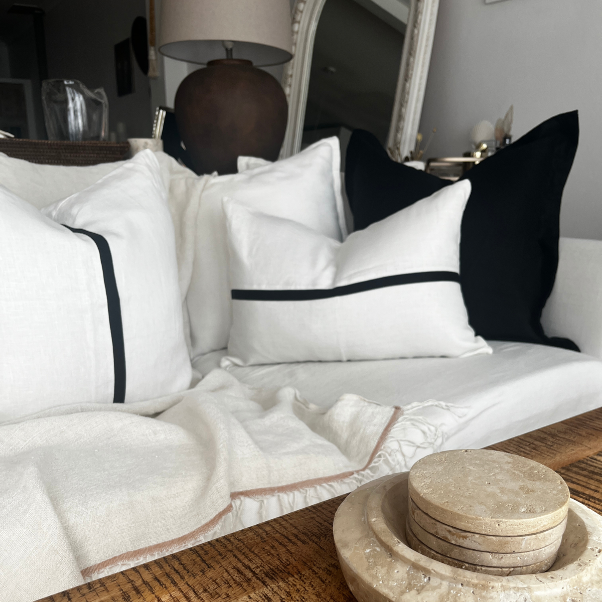White linen throw pillows with black trim accent arranged on a white sofa alongside a solid black accent pillow and throw blanket.