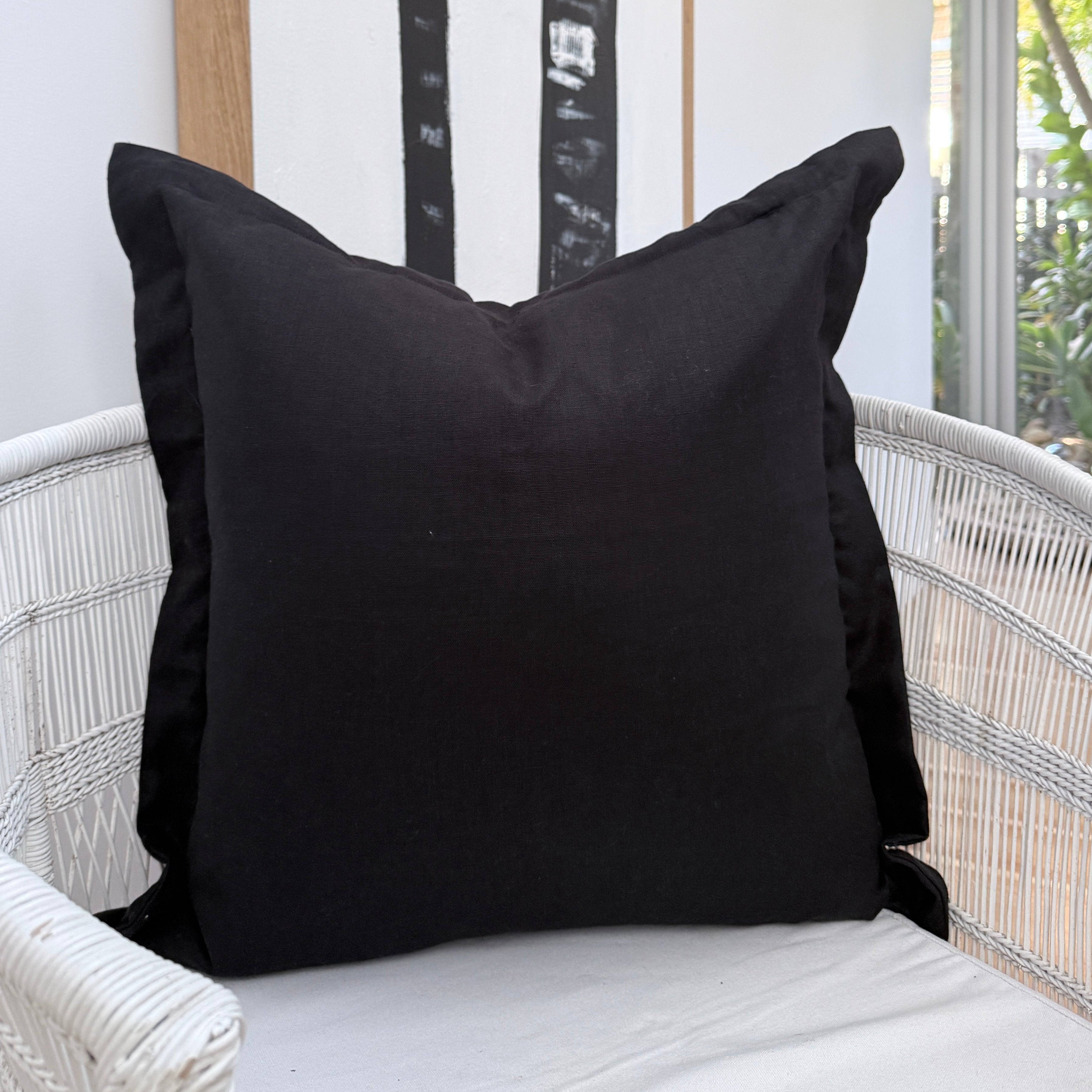 A black square throw cushion with a ruffled edge resting on a white woven outdoor chair, with a white wall and a framed black-and-white palm tree print in the background.