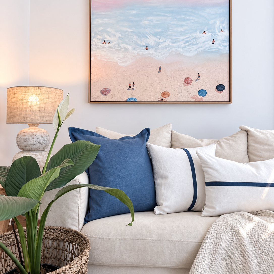 Blue and white linen cushion arrangement styled on a white couch beneath beach artwork.
