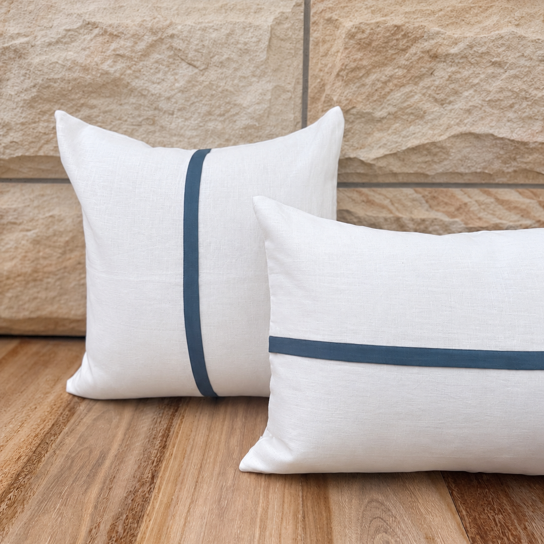 Two white linen throw pillows with navy blue stripe accents, styled on a wooden floor against a stone wall background.