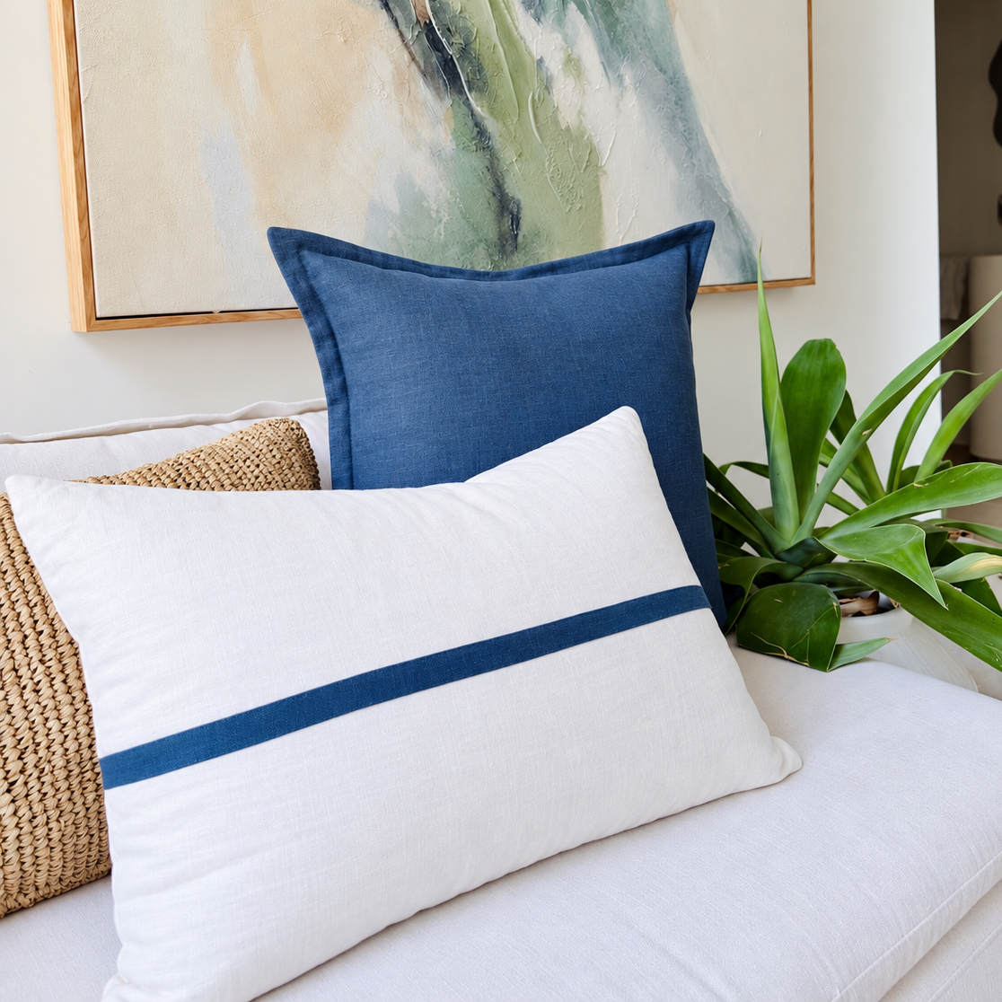 Blue solid and white blue-striped linen cushions styled on a sofa next to a green potted plant.