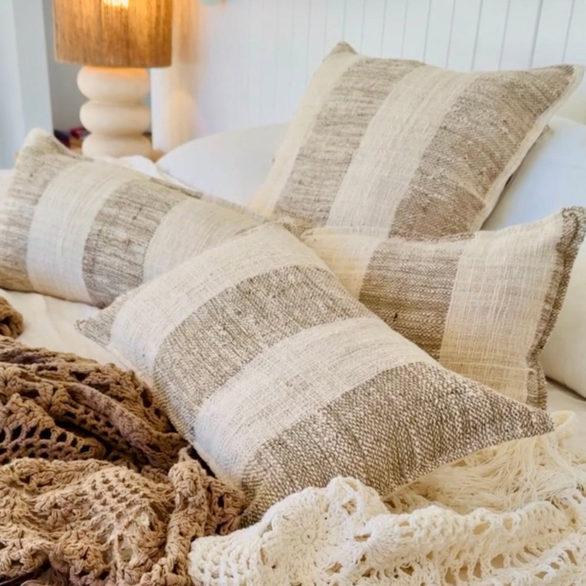 Three woven throw pillows with beige and brown horizontal stripes arranged on a bed with a cream crocheted blanket and a warm lamp in the background.