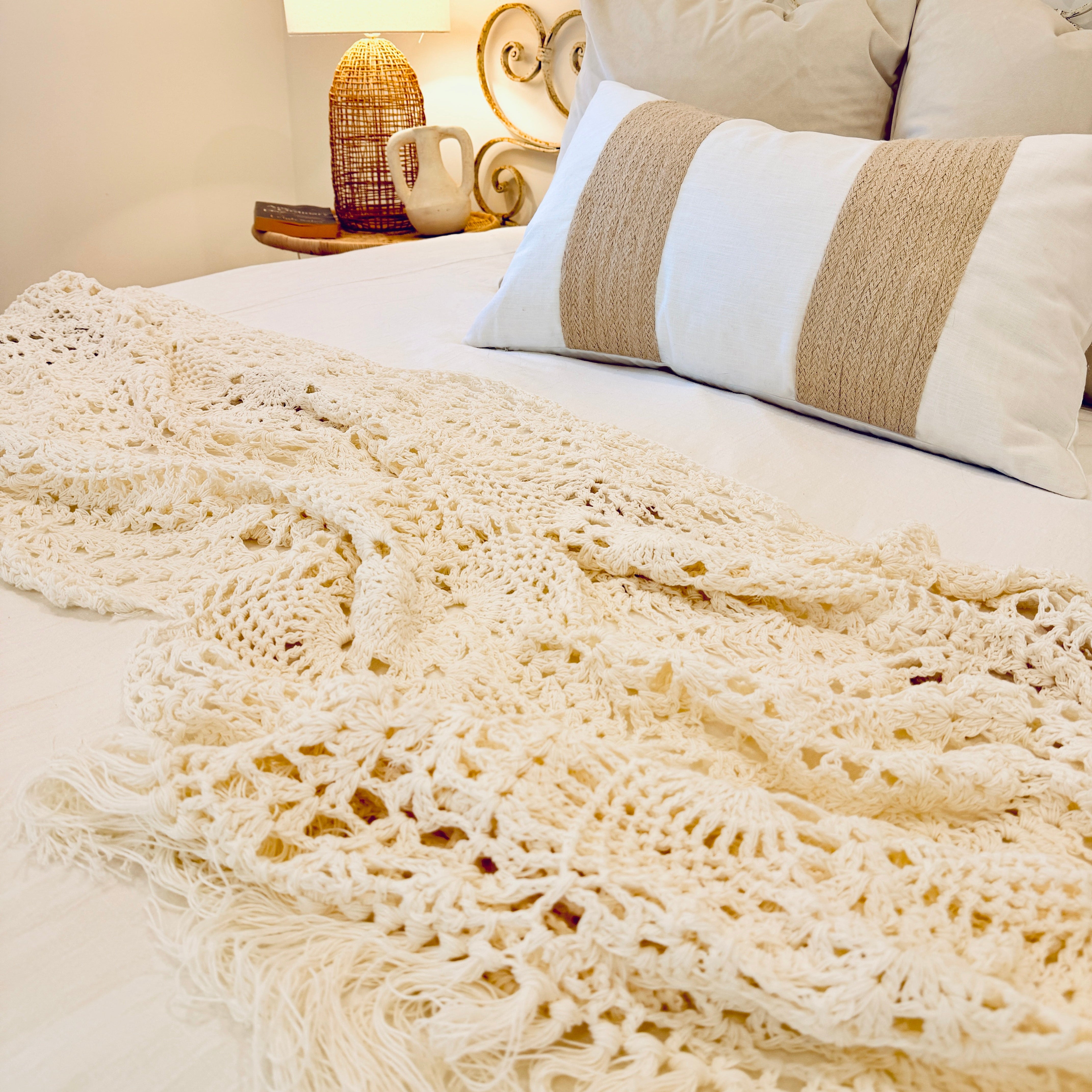 Cozy bedroom with a bed featuring a crochet blanket, cushions, and a nightstand with a lamp.