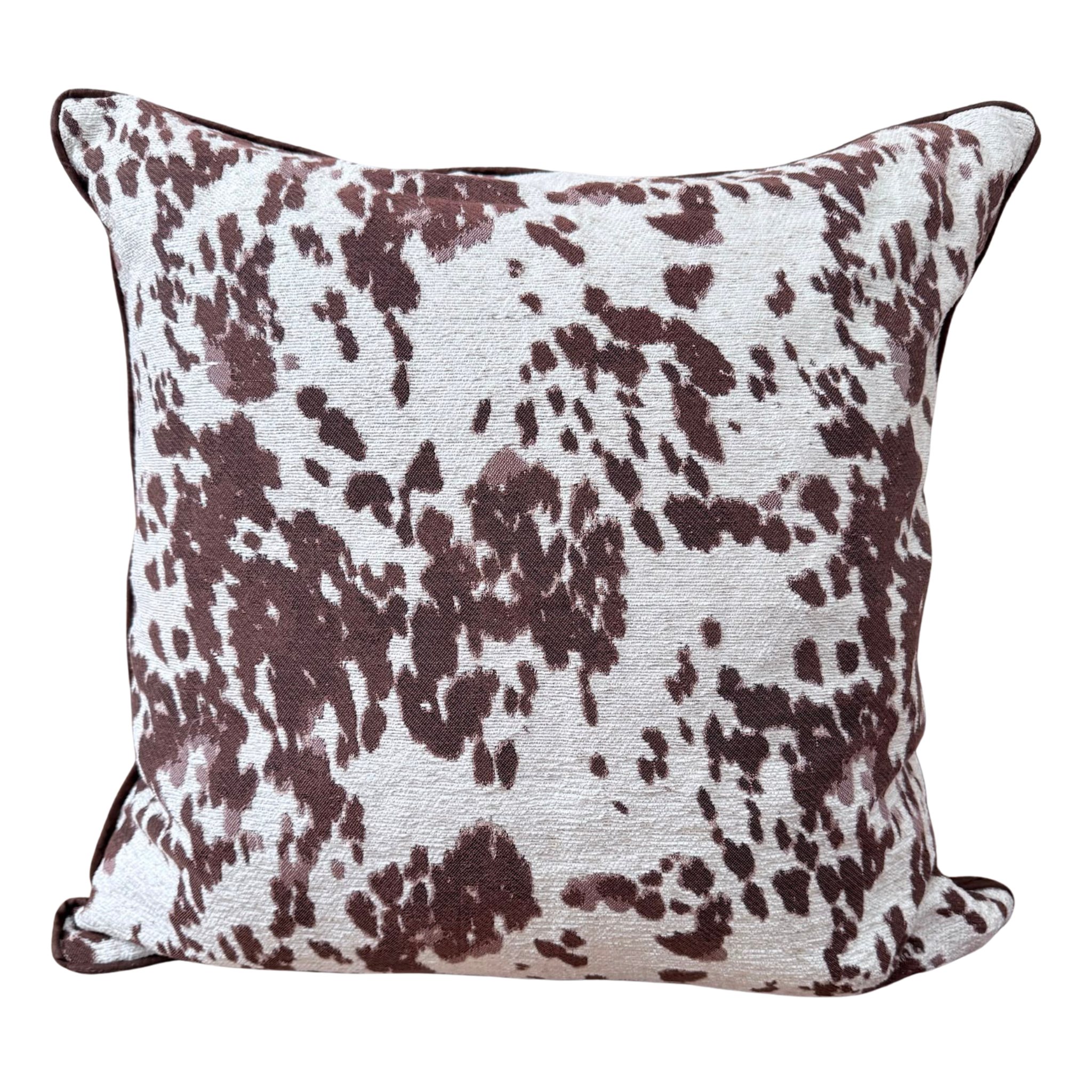 A square throw cushion featuring a brown and white cow-print pattern with dark brown piped edges, shown against a plain white background.