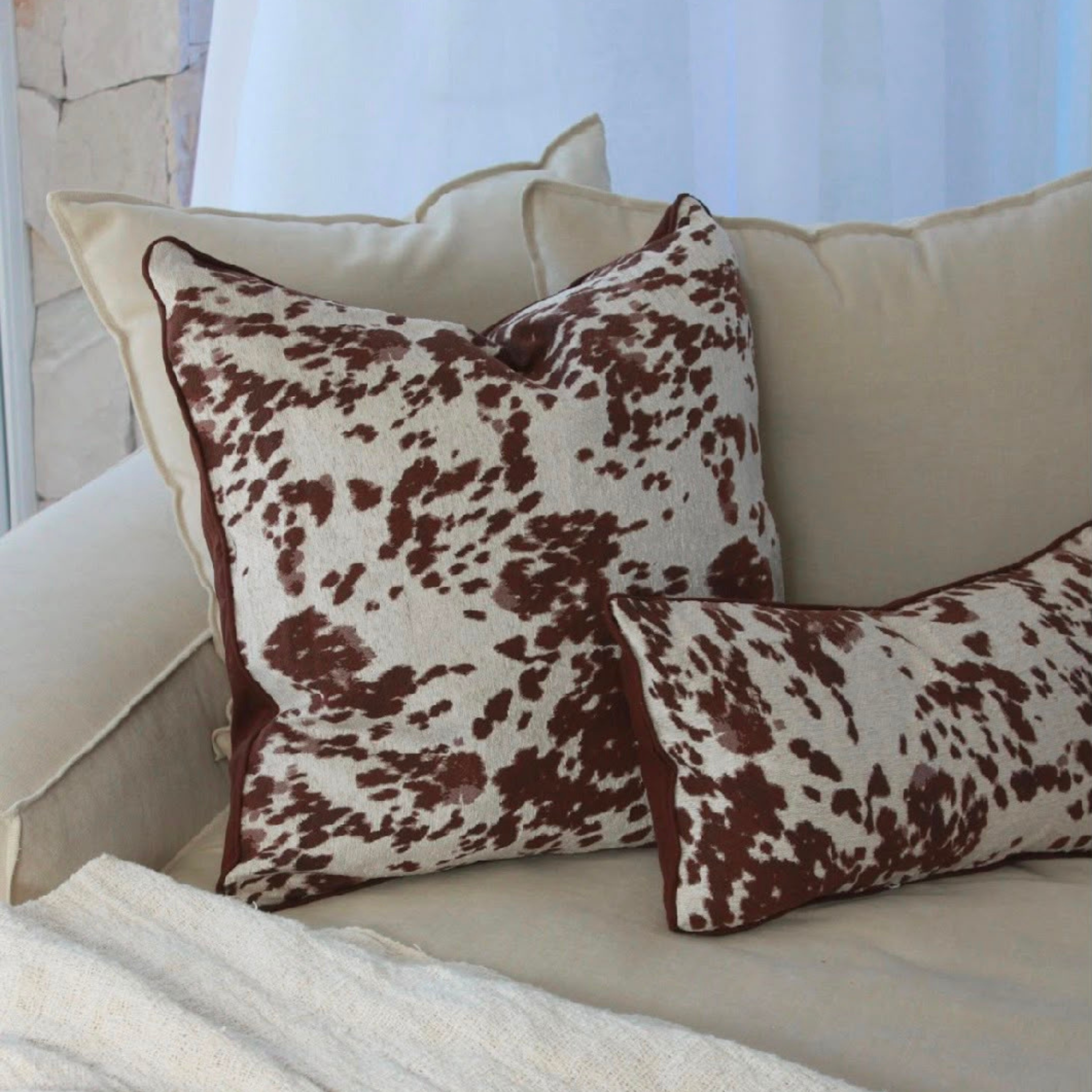 Two decorative throw cushions with a brown and cream cow print pattern, styled on a beige sofa with a neutral background.