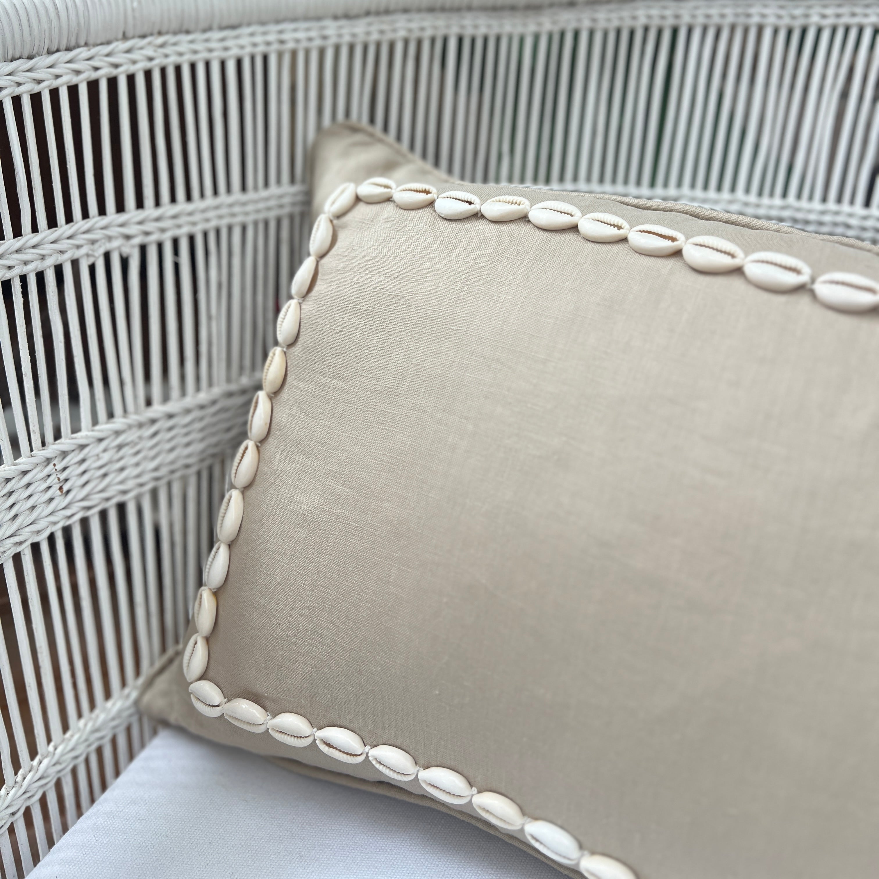 A beige linen cushion with a rectangular pattern made from sewn on shells, displayed in a wicker chair.