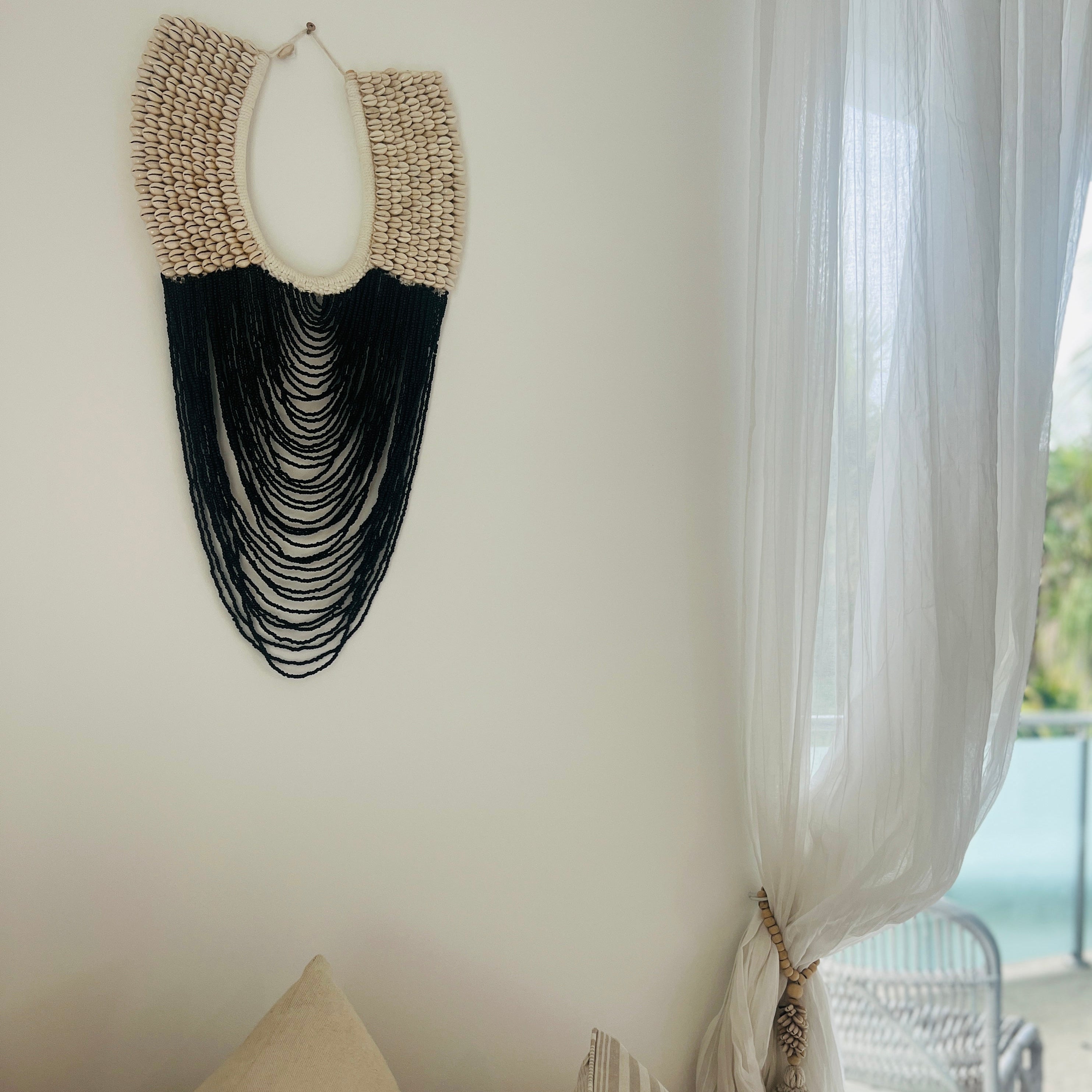 A wall-mounted cowrie shell necklace with black beading, hanging above a bed in a room with light decor.