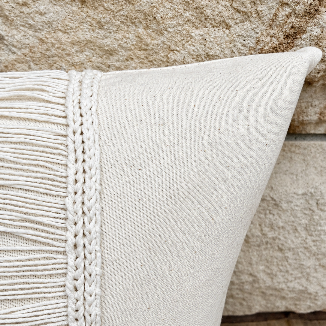 Corner detail view of a cream linen pillow showing smooth edge seam and braided fringe border.