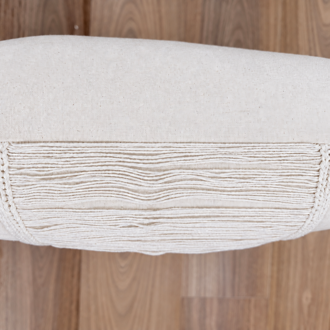 Top-down view of cream linen cushion showing central fringe overlay on a wooden surface.