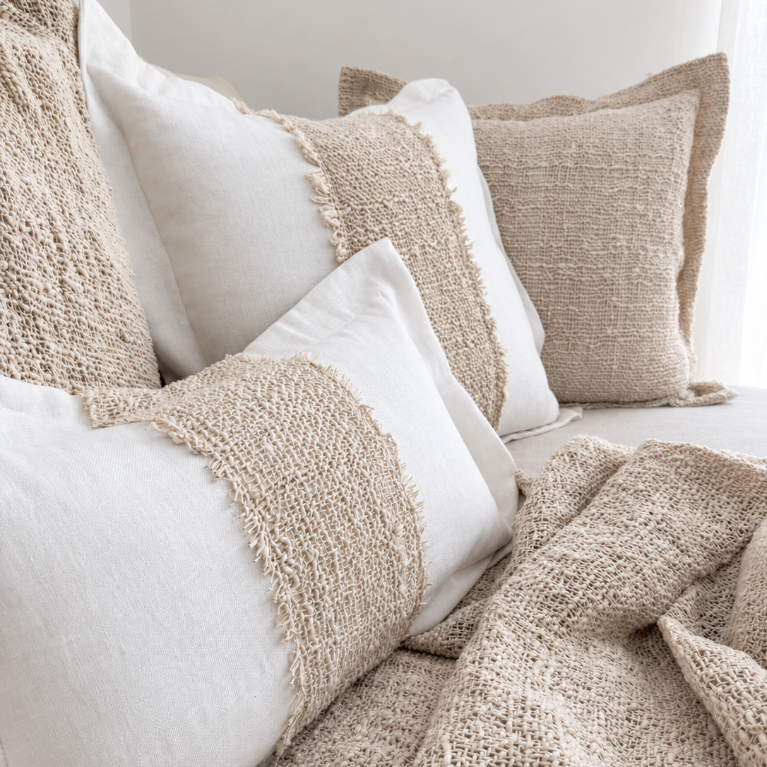 A cozy arrangement of neutral-toned  pillows with chunky woven jute-style bands, a matching beige throw blanket, and a soft neutral throw on a bed.