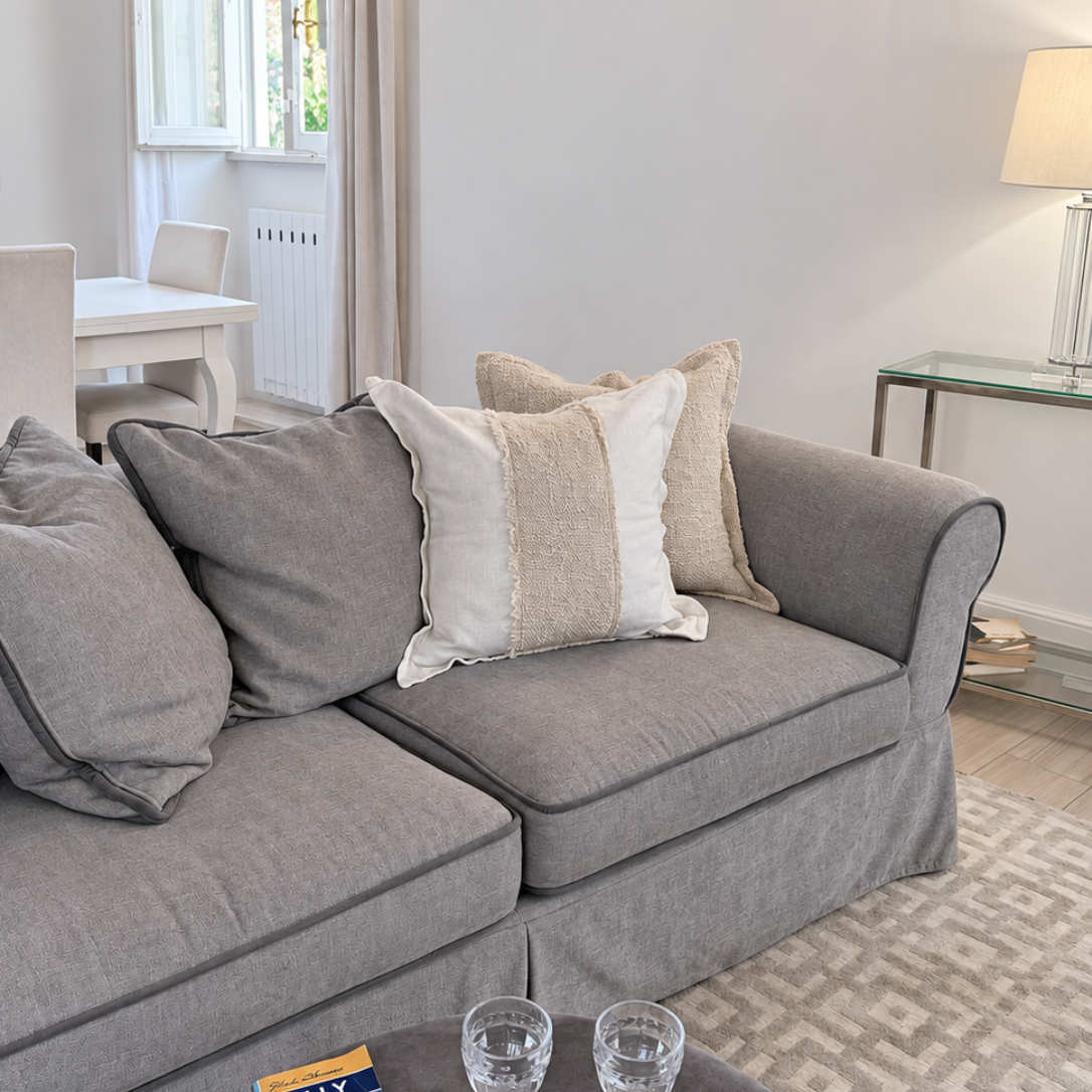 Square white linen throw cushion with a textured woven center stripe styled on a grey sofa.