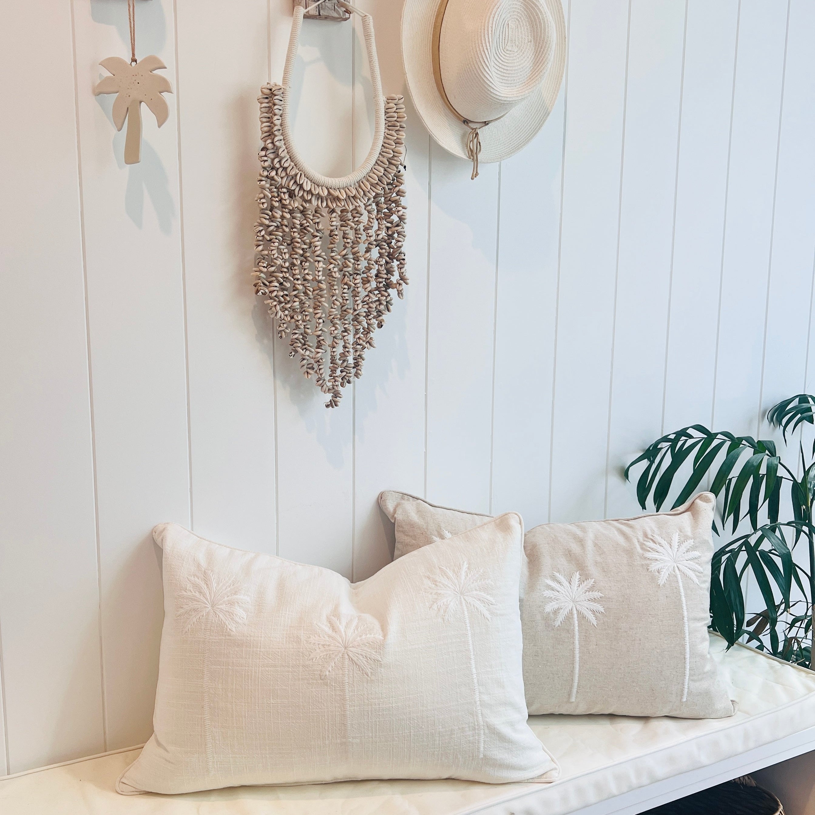 Two cushion covers with embroidered palm trees and a cowrie shell hanging on a white wall shelf, with decorative items in the background.