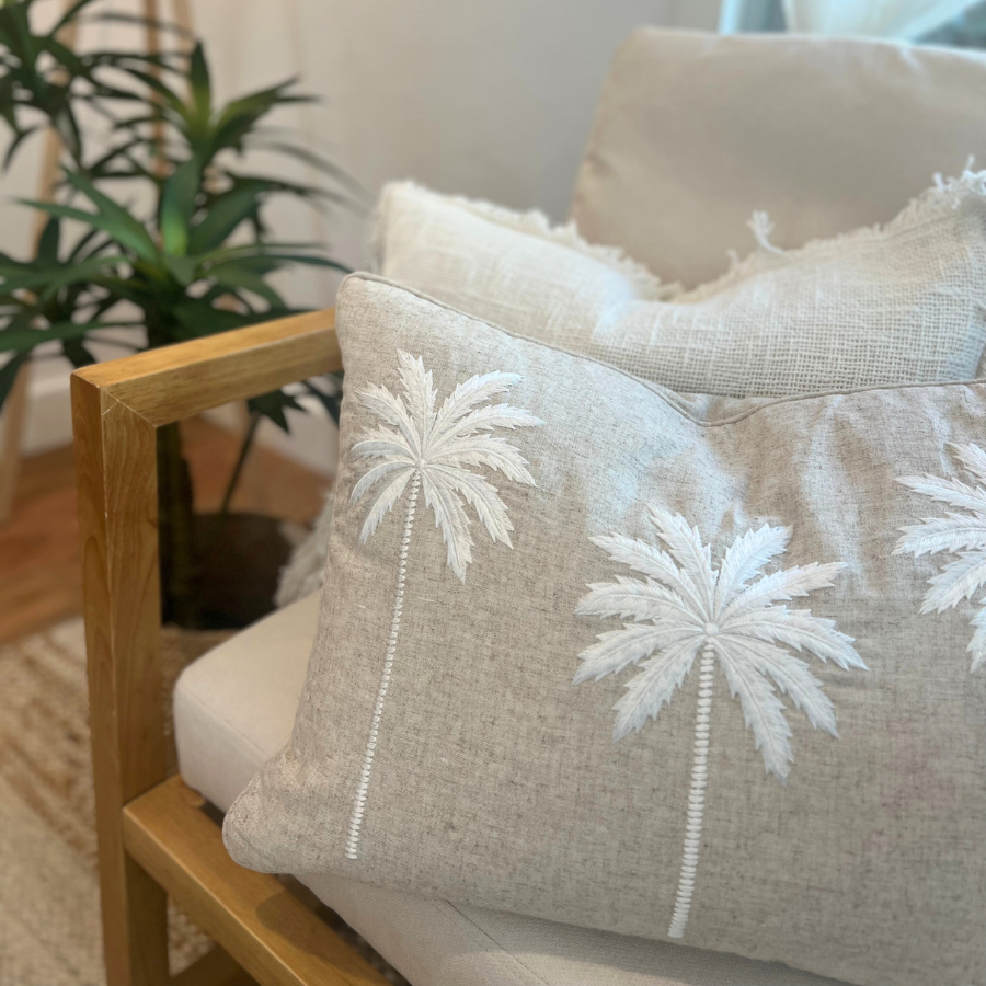 Two cushions with palm tree decor on a chair with plant and lamp.