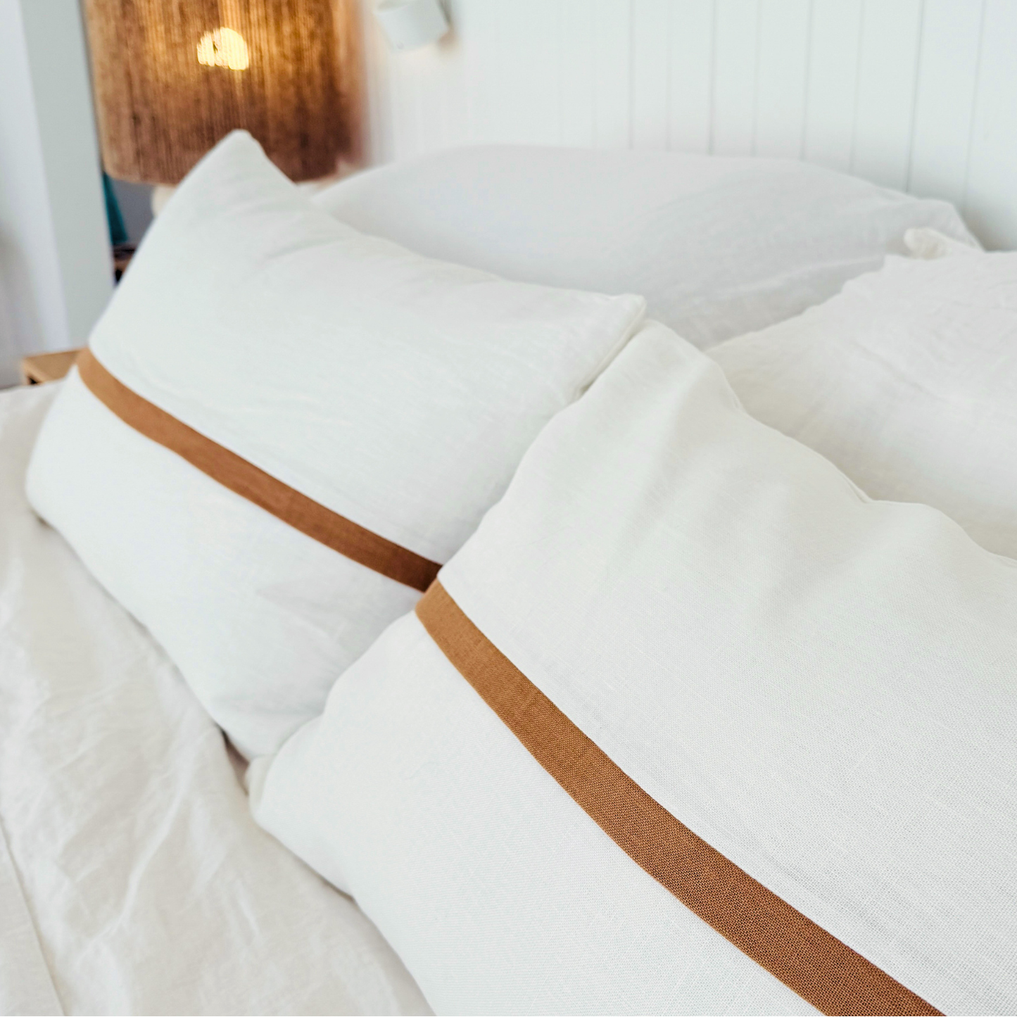 White cushion with brown trim on a bed in a softly lit bedroom.