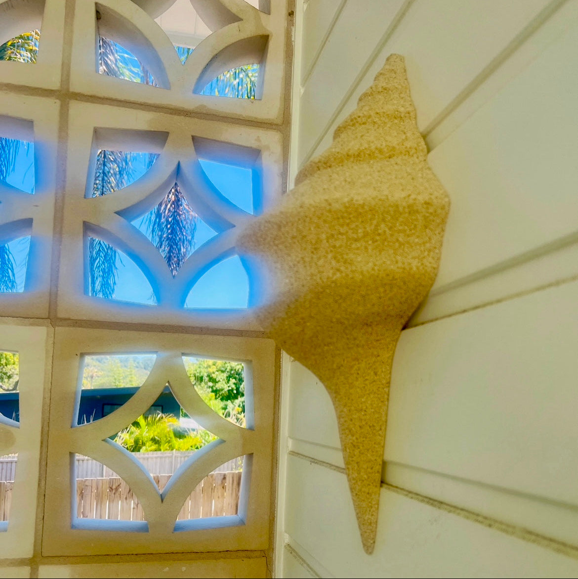 A conch shell-shaped wall decor in a natural sand finish, mounted on an outdoor wall with lattice work and greenery in the background.