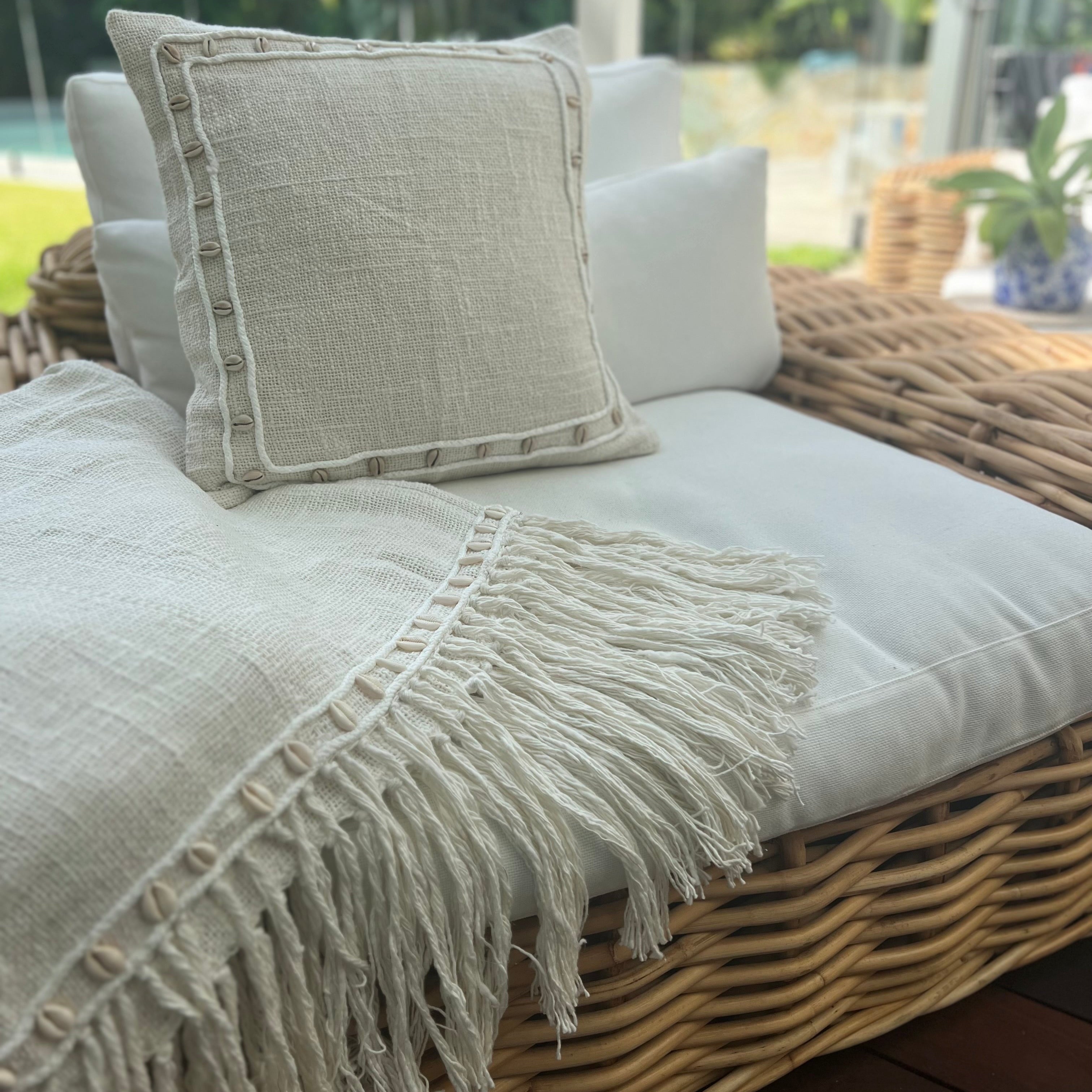 A beige cotton cushion cover with hand-sewn cowrie shells and fine rope stitching, accompanied by a matching throw with fringing at the ends, displayed on a wicker sofa.